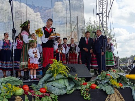 Zdjęcie przedstawia obrzęd przekazania bochna chleba włodarzom gminy. W tle Zespól Promni