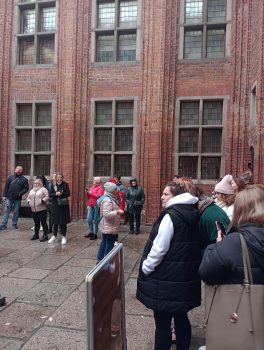 Zdjęcie przedstawia grupę znajdującą się  na dziedzińcu, skierowaną ku stojącemu na środku przewodnikowi. 