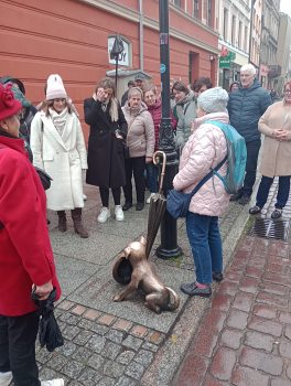 Zdjęcie przedstawia  zwiedzających skierowanych ku przewodnikowi oraz do pomnika przedstawiającego psa z kapeluszem i parasolką. 