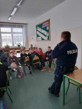 Na zdjęciu na pierwszym planie widzimy policjanta opartego tyłem o ławkę ,a w tle siedzące  na krzesłach  dzieci