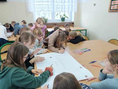 Na zdjęciu widzimy grupkę dzieci siedzących przy ławkach i malujących flamastrami na dużym arkuszu papieru plakat