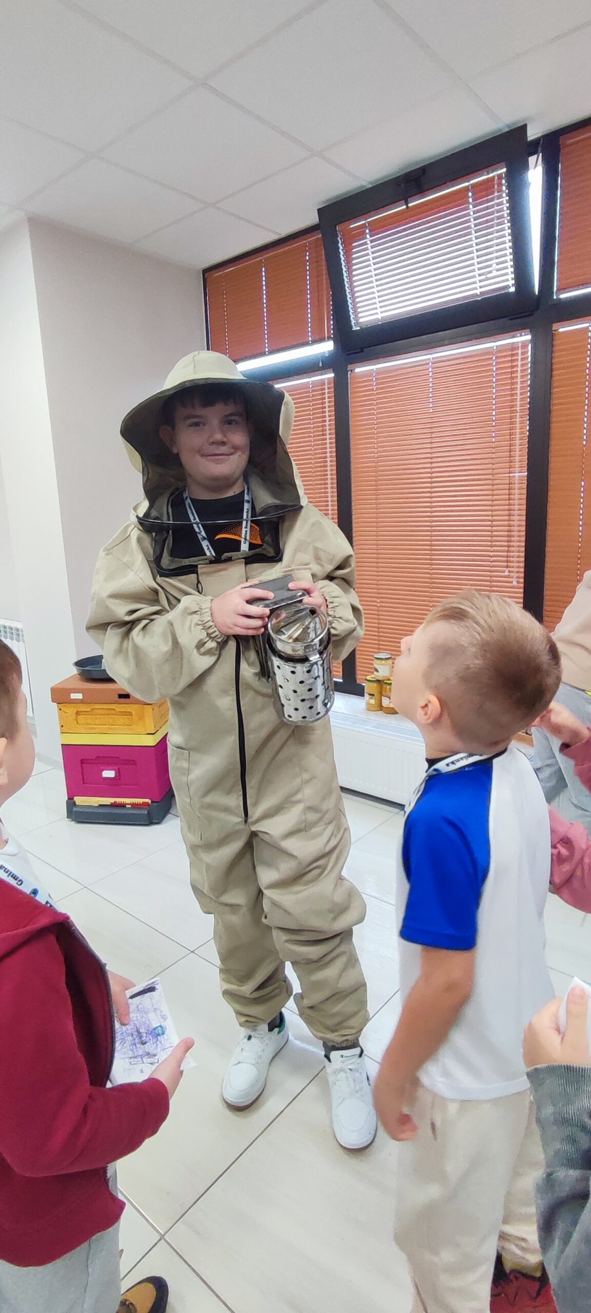 Zdjęcie przedstawia chłopca przebranego w strój pszczelarza z dymiarką w ręku, wokół inni zainteresowani przymierzeniem stroju.