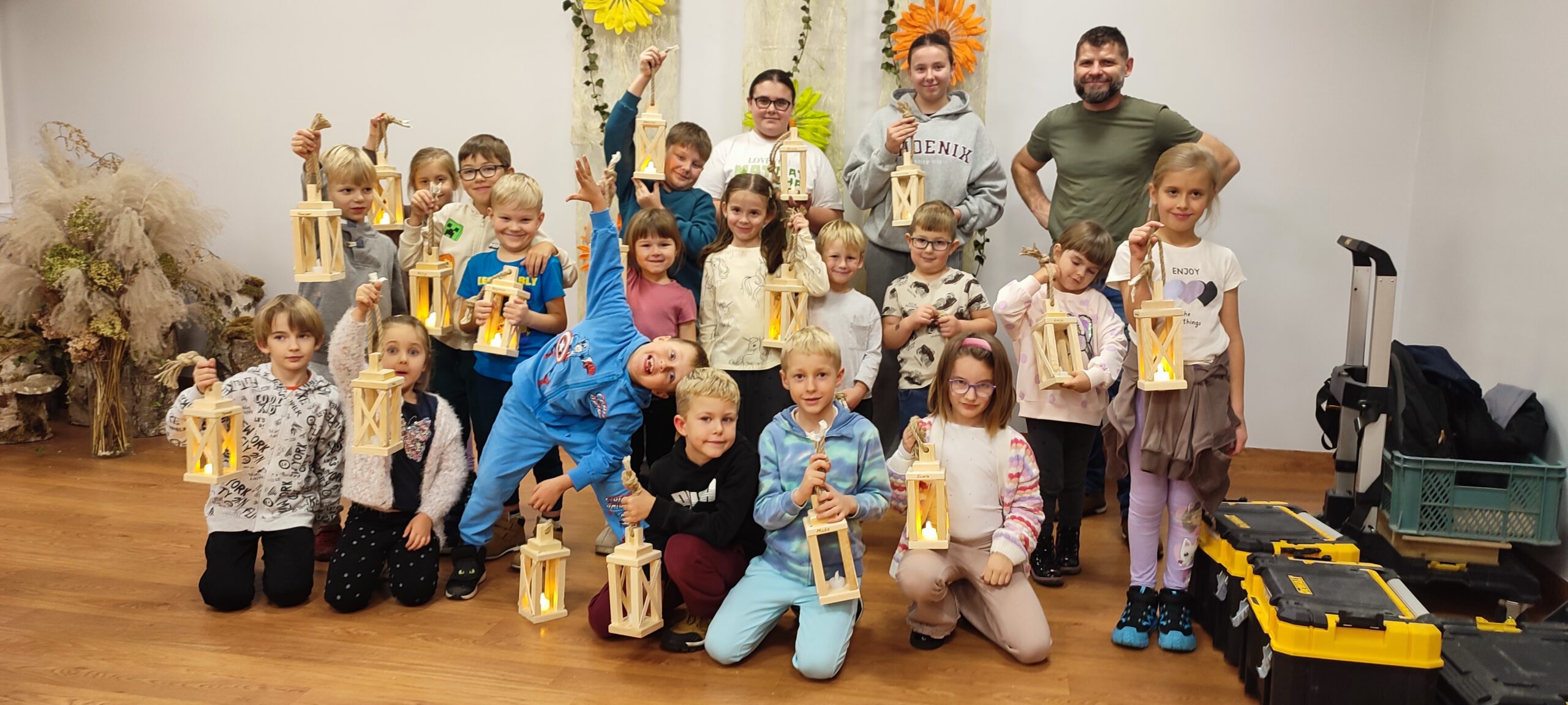 Na zdjęciu widać dużą, radosną grupę dzieci pozujących w pomieszczeniu wraz z panem prowadzącym warsztaty majsterkowania. Wszystkie dzieci trzymają w dłoniach drewniane lampiony z żółtym, ciepłym światłem w środku — własnoręcznie wykonane podczas warsztatów. Grupa stoi i klęczy na podłodze, uśmiechając się do aparatu. W tle widać dekoracje z suszonych roślin, dużych kwiatów i materiałowych ozdób, co nadaje całości przyjemny, warsztatowo-artystyczny klimat. Po prawej stronie znajdują się skrzynki i narzędzia, po warsztatach z majsterkowania.