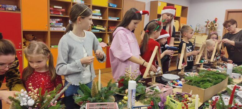 Na zdjęciu widać świąteczne warsztaty stroików bożonarodzeniowych, w których biorą udział dzieci wraz z paniami z KGW Nowe Kozłowo. Atmosfera jest radosna i zimowo-świąteczna. Dzieci i dorośli pracują wspólnie przy długich stołach, na których rozłożone są gałązki świerkowe, ozdoby choinkowe, bombki, szyszki, świece, wstążki oraz inne materiały dekoracyjne. Uczestnicy tworzą dwa rodzaje dekoracji: stroiki świąteczne z zielonych gałązek i świec, drewniane trójkątne „choinki”, które wypełniają bombkami i ozdobami. Kilka osób korzysta z kleju na gorąco, aby przymocować elementy dekoracyjne. W tle znajdują się regały z zabawkami i grami, co sugeruje, że akcja odbywa się w sali GOK Somianka.