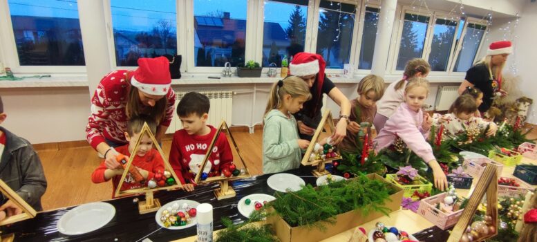 Zdjęcie przedstawia dzieci i dorosłych pracujących wspólnie przy długich stołach, na których rozłożone są gałązki świerkowe, ozdoby choinkowe, bombki, szyszki, świece, wstążki oraz inne materiały dekoracyjne. Uczestnicy tworzą dwa rodzaje dekoracji: stroiki świąteczne z zielonych gałązek i świec, drewniane trójkątne „choinki”, które wypełniają bombkami i ozdobami. Kilka osób korzysta z kleju na gorąco, aby przymocować elementy dekoracyjne. Część dzieci i dorosłych ma na głowach świąteczne nakrycia, takie jak czapki Mikołaja czy opaski z rogami renifera. W tle widać duże okna udekorowane lampkami, za którymi jest już ciemno — podkreśla to zimowy, przedświąteczny klimat.