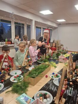 Zdjęcie przedstawia dzieci i dorosłych podczas prac przy stroikach bożonarodzeniowych. Część dzieci i dorosłych ma na głowach świąteczne nakrycia, takie jak czapki Mikołaja czy opaski z rogami renifera. W tle widać duże okna udekorowane lampkami, za którymi jest już ciemno — podkreśla to zimowy, przedświąteczny klimat.