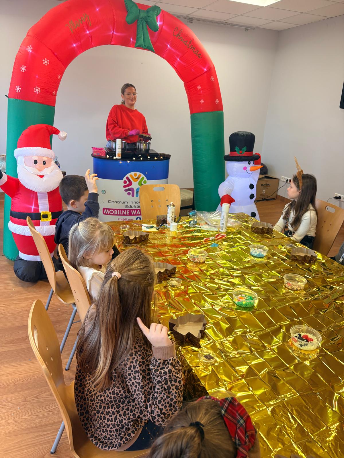 Zdjęcie przedstawia świąteczne warsztaty dla dzieci. Przy stole przykrytym złotą, błyszczącą folią siedzi grupa dzieci, a przed nimi stoją miseczki z kolorowymi dodatkami i ozdobami. Z przodu widoczne są nadmuchiwane dekoracje – Mikołaj oraz bałwan. Pod łukiem świątecznym stoi kobieta w czerwonym swetrze, która prowadzi zajęcia i podgrzewa czekoladę. Na tym zdjęciu troje chłopców siedzi przy stole pokrytym złotą, połyskującą folią. Przed każdym z nich stoją miseczki z kolorowymi ozdobami do dekorowania. Chłopiec pośrodku skupia się na swojej pracy, dekorując brązową formę w kształcie choinki. Dwaj pozostali patrzą w stronę aparatu lub w jego kierunku. W tle widać kolorowe szafki z zabawkami. Dwóch chłopców siedzi przy stole przykrytym złotą, połyskującą folią. Pracują nad świątecznymi, choinkowymi formami do czekolady. Jeden chłopiec w czerwonej koszuli trzyma pustą foremkę w kształcie choinki, a drugi, w swetrze z zimowym wzorem, nakłada słodkie dodatki do swojej foremki. Na stole widoczne są małe miseczki z dodatkami.