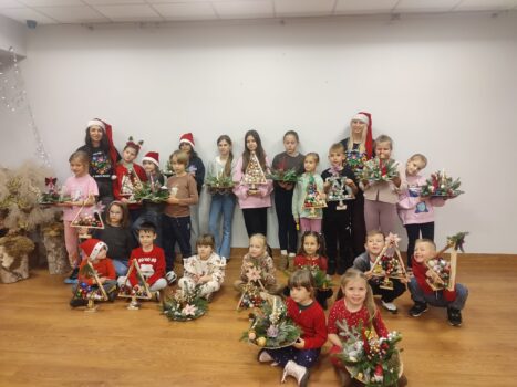 Na zdjęciu widać grupę dzieci i dwie dorosłe osoby pozujące razem w Sali po przeprowadzonych warsztatach świątecznych. Wszyscy trzymają w rękach własnoręcznie wykonane dekoracje bożonarodzeniowe – stroiki, małe choinki. Większość dzieci siedzi lub klęczy z przodu, a reszta stoi z tyłu. Dwie dorosłe osoby stoją po bokach i mają na głowach czerwone czapki Mikołaja. Wiele dzieci również ma świąteczne ubrania lub akcesoria, np. mikołajkowe czapki czy swetry. Cała scena wygląda bardzo radośnie i świątecznie – to grupowe zdjęcie z zakończenia kreatywnych warsztatów bożonarodzeniowych.